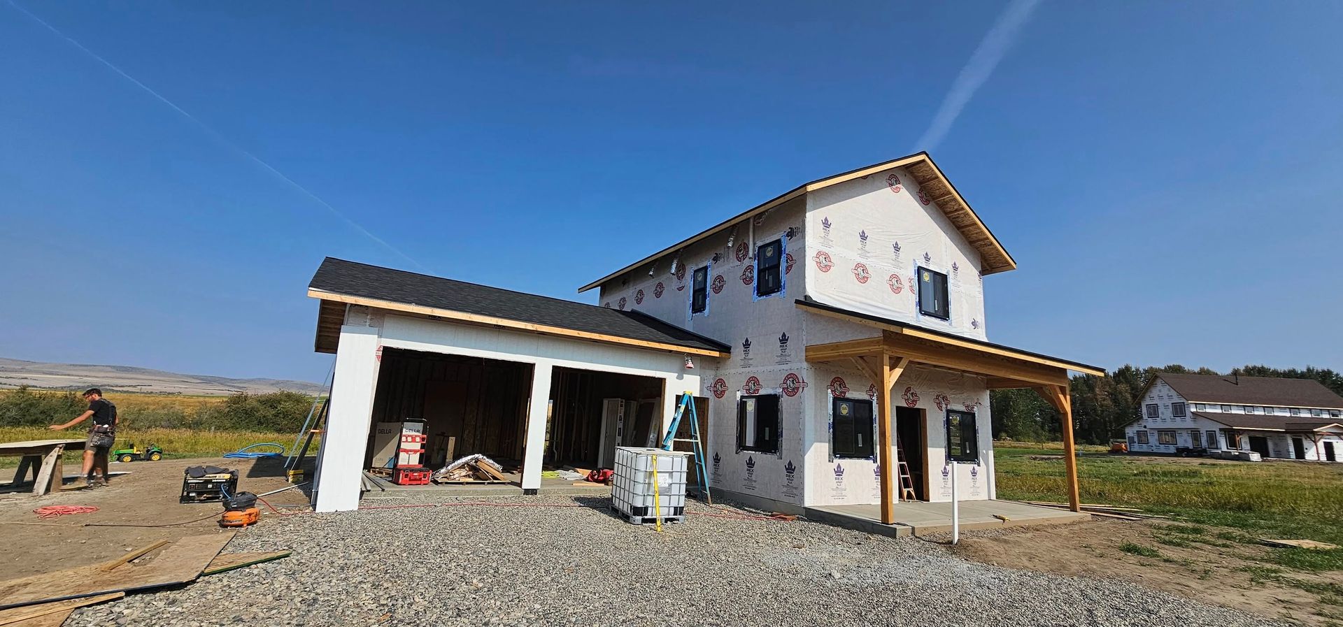 A white two-story house under construction with an attached garage, featuring exposed wood framing and a gravel driveway.