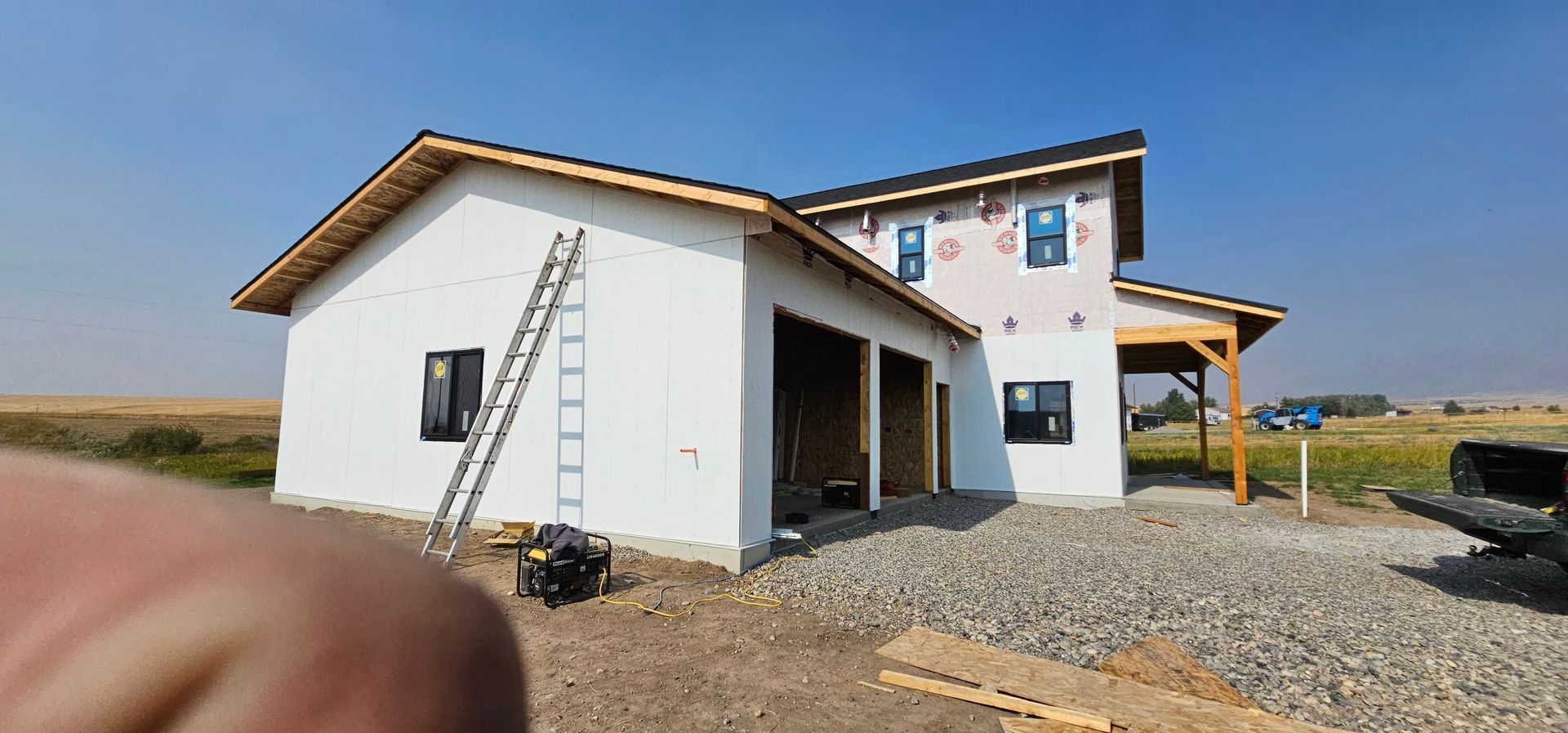 An unfinished, single-story house with white exterior walls, a dark roof, and open garage bays sits in a rural field.