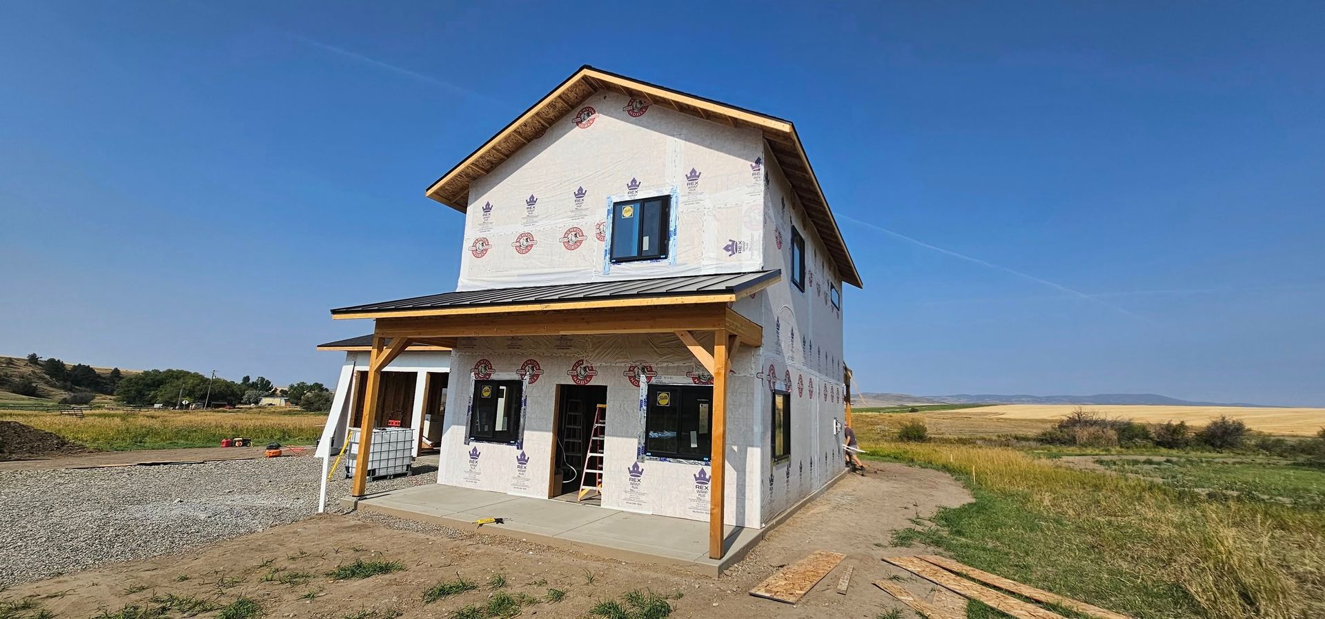 A two-story house under construction with light siding and a wooden porch, set in a rural, open landscape under blue sky.