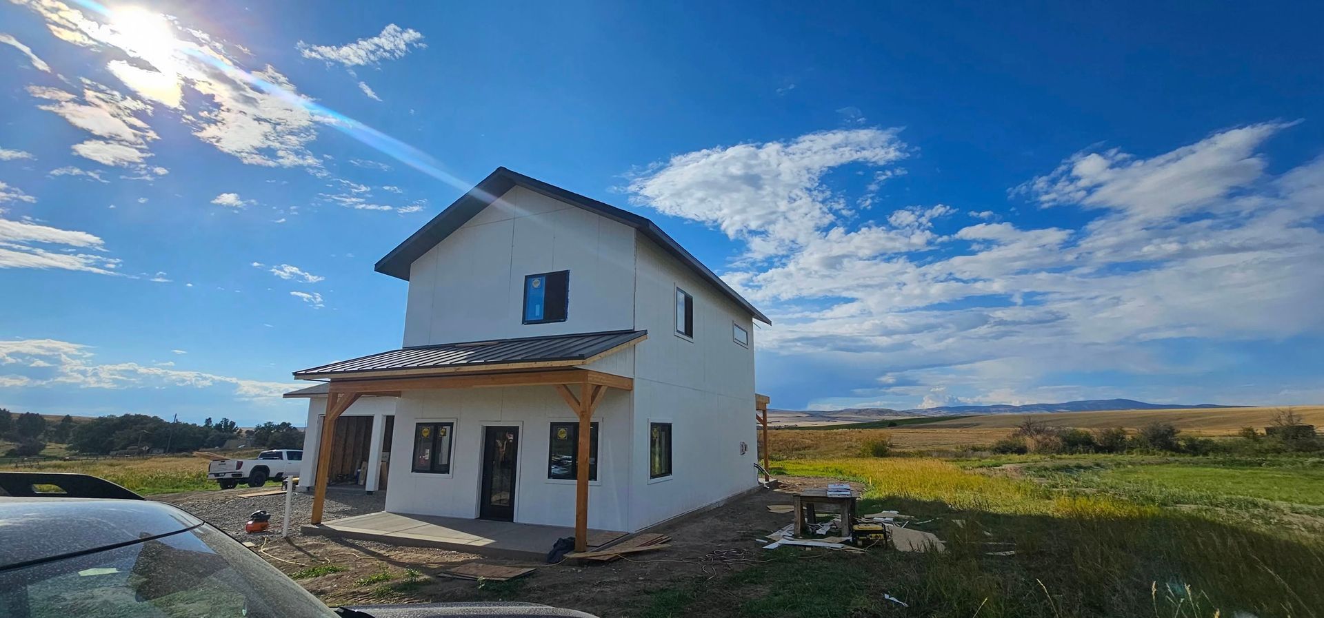 A white two-story house with a wooden porch sits in a sunny field under a bright blue sky with scattered clouds.