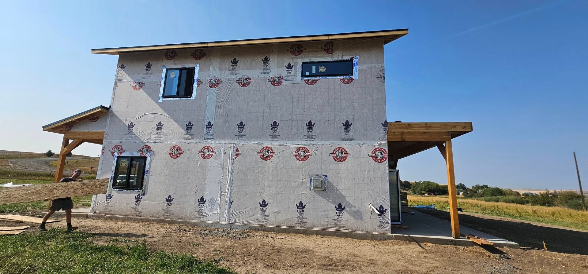 Two-story house under construction with exposed exterior insulation board, installed windows, and wooden porch framing.