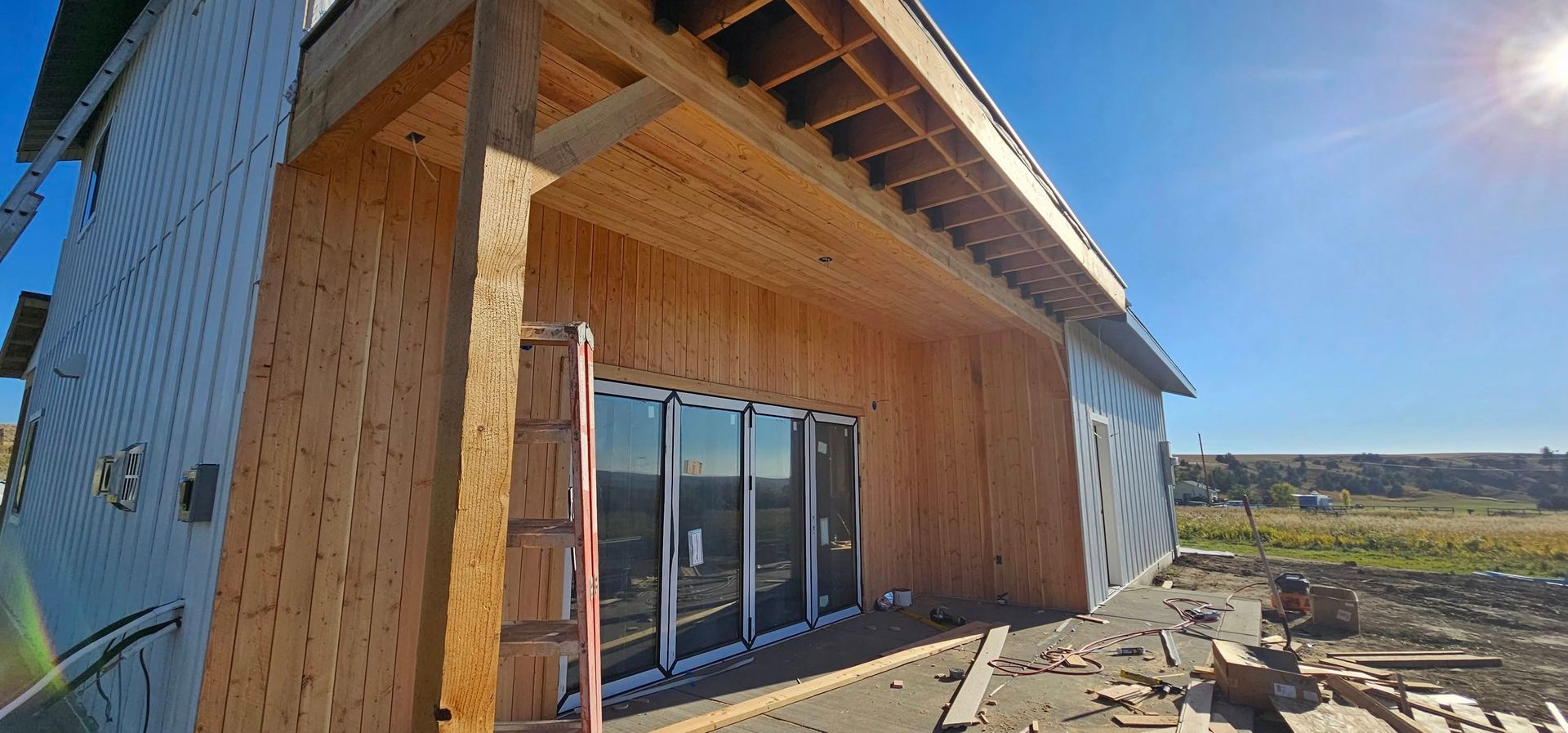A house under construction with exposed wooden framing and siding, featuring a patio door area under a porch overhang.