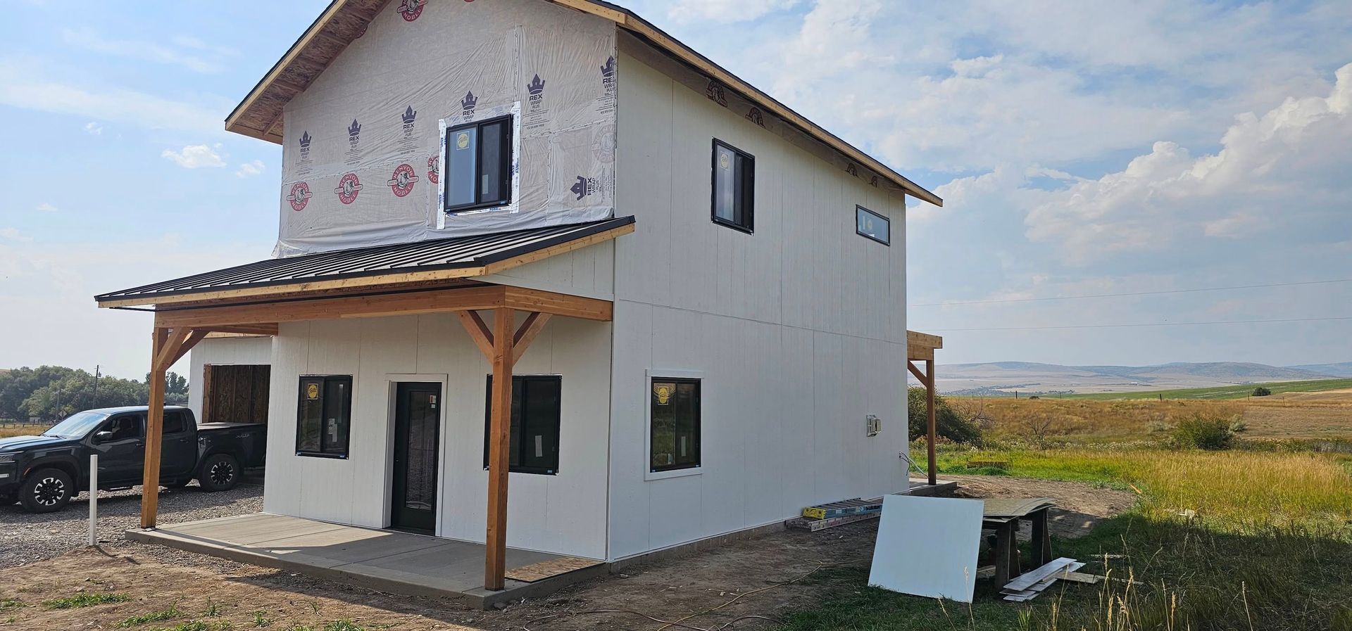 A two-story house under construction with a wooden porch and white siding in a rural field under a blue sky.