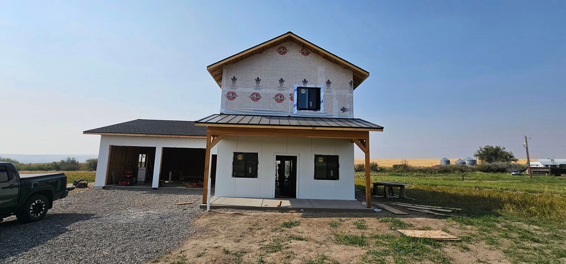 A two-story house under construction with white stucco walls, a dark shingled roof, and a gravel driveway in a rural field.