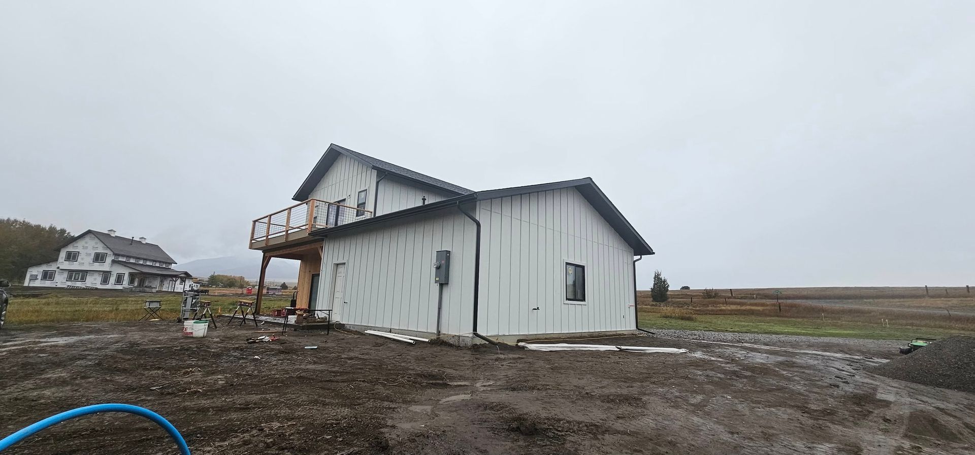 A white two-story house under construction with an unfinished exterior, wooden deck, and a muddy yard on a cloudy day.
