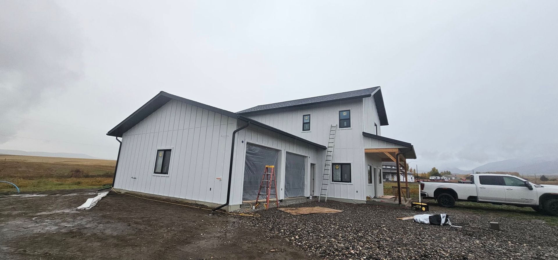 A newly constructed two-story house with white exterior siding stands on a gravel lot under a cloudy, overcast sky.