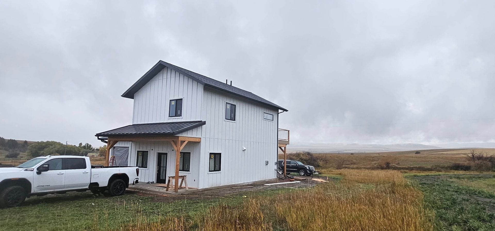 A two-story white house with a covered porch and a white truck parked in front, situated in a field under cloudy skies.