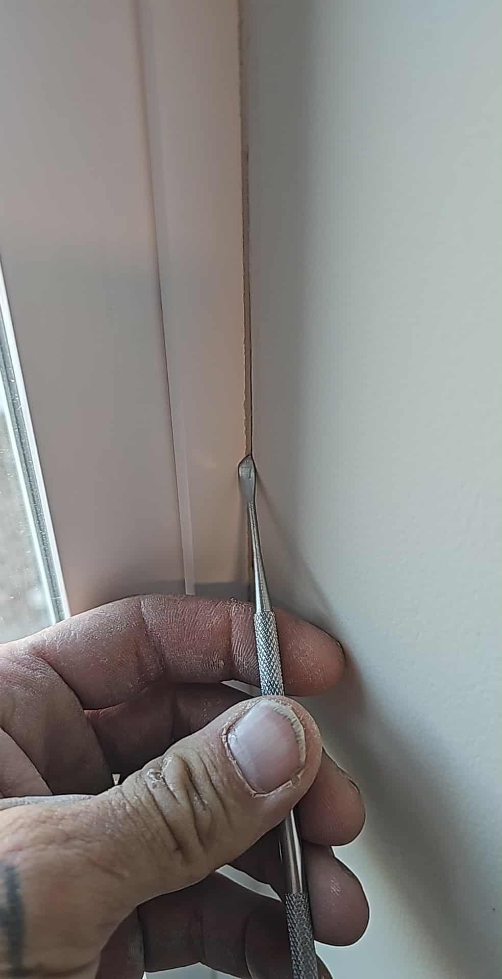 A close-up of a hand using a metal tool to peel back a strip of sealant from the edge of a white window frame.