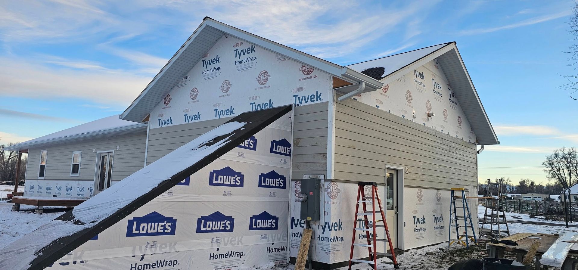 A single-story house under construction with white exterior sheathing, partial beige siding, and ladders in snowy weather.