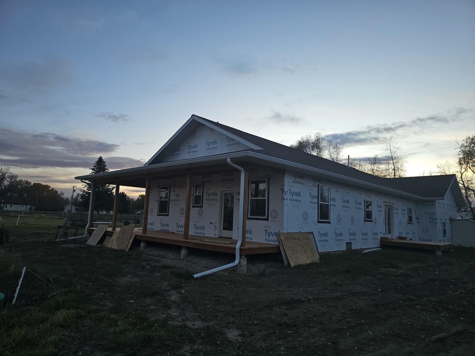 A house under construction with a front porch, white house wrap, and exposed wooden frame at twilight.