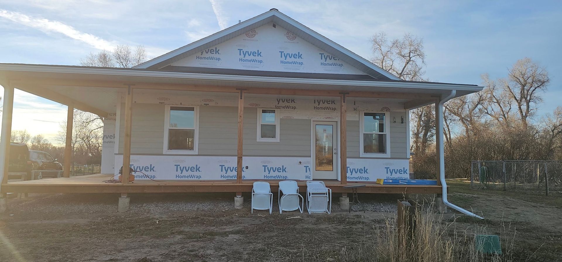 A single-story house under construction with white Tyvek house wrap, partially installed siding, and a wooden front porch.