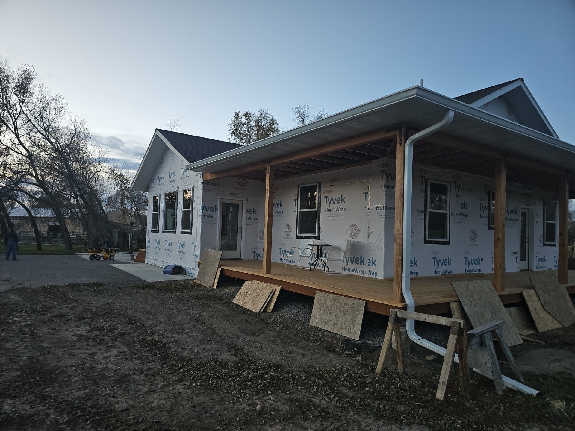 A house under construction with white exterior sheathing, a wooden porch, and a white downspout on a dirt lot.