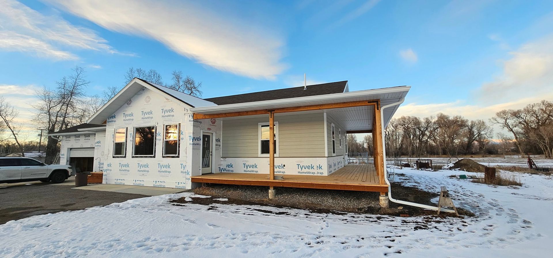 A house under construction with white exterior sheathing, a large wooden front porch, and snow-covered ground.
