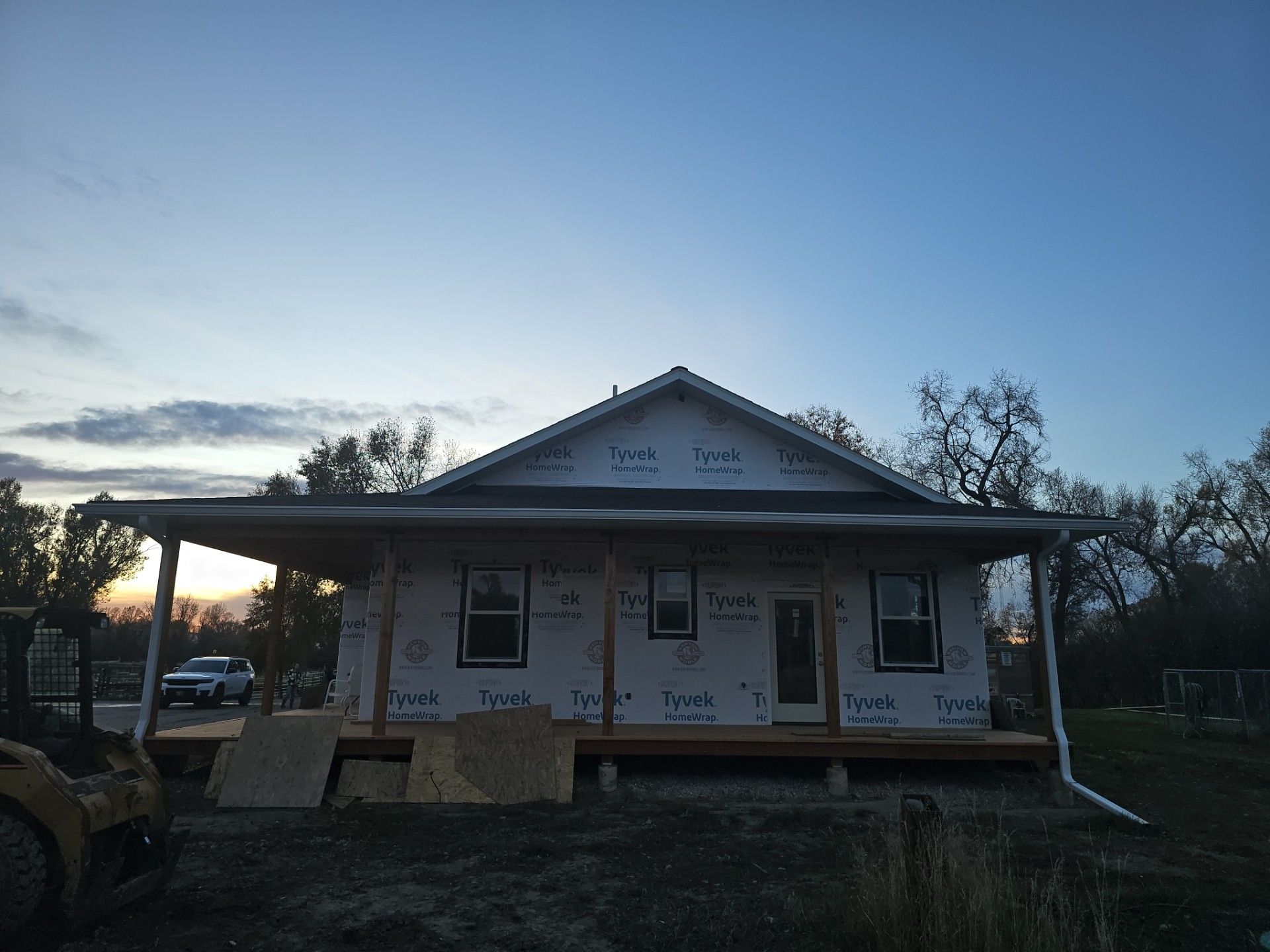 A house under construction with a front porch, white weather-resistant wrap on the walls, and a gabled roof at sunset.