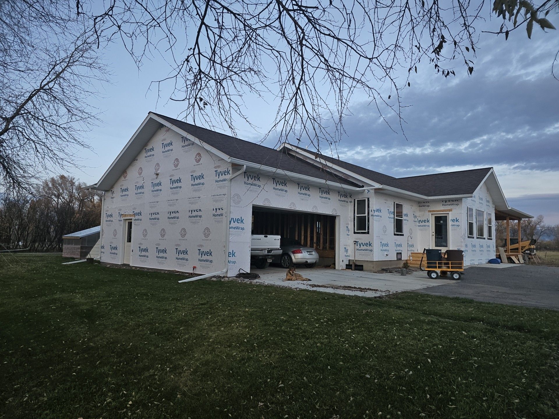 A house under construction with a garage, covered in white house wrap, set on a lawn at dusk.