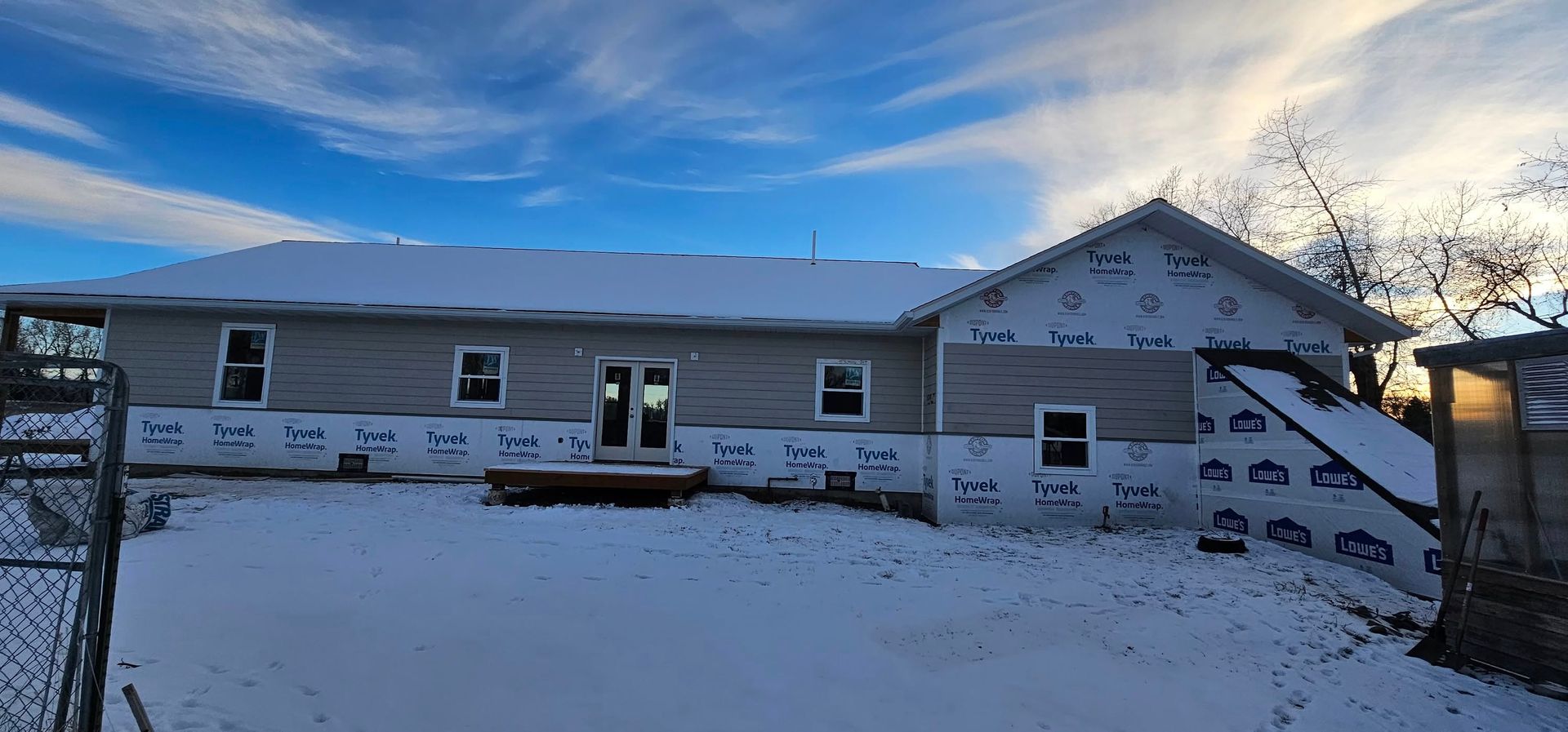A single-story house under construction with light gray siding and white house wrap, set against a snowy yard and blue sky.
