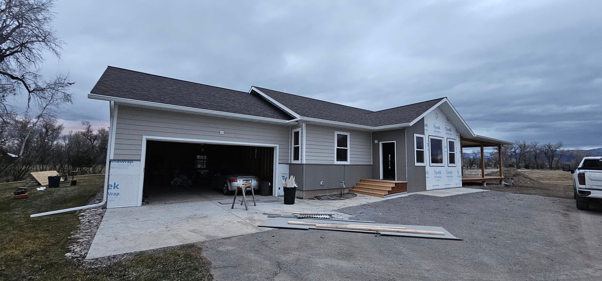 A newly constructed house with a garage, light siding, a dark shingled roof, and an unfinished gravel driveway.