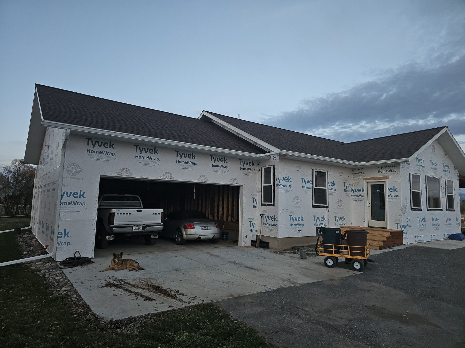 A partially constructed house with white exterior insulation, dark roofing, and a two-car garage containing two vehicles.