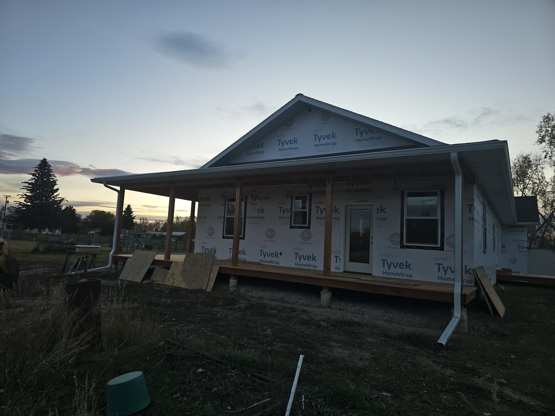 A house under construction with white Tyvek house wrap, a wooden porch, and installed windows at twilight.