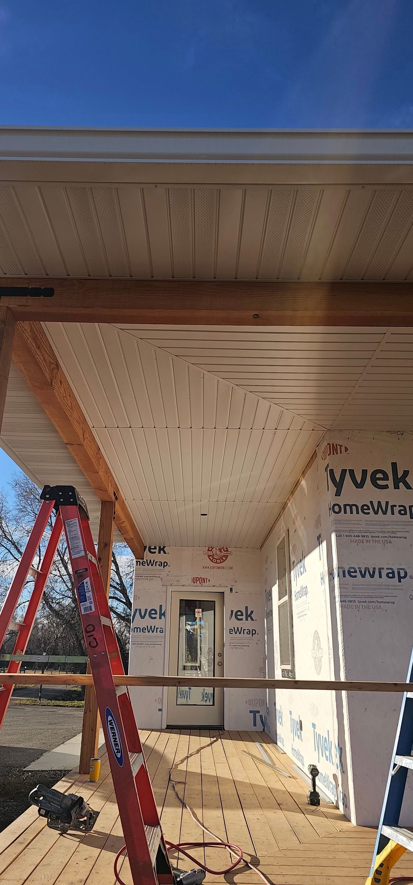 A low-angle view of a house porch under construction, featuring white perforated soffits, Tyvek siding, and wooden decking.