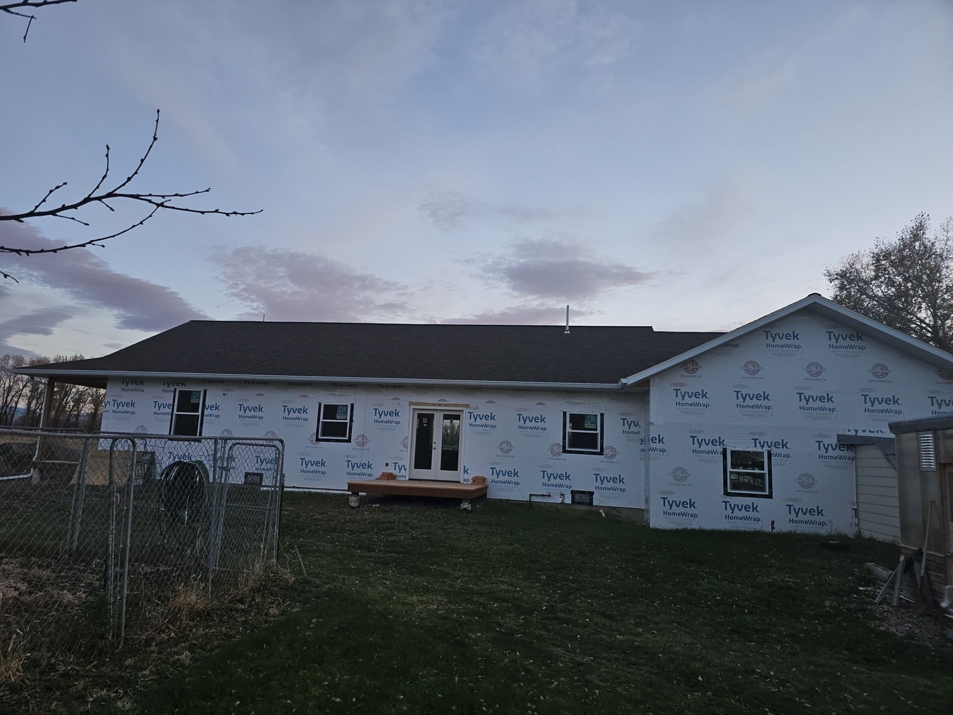 A single-story house under construction with white exterior house wrap, dark roof shingles, and installed windows.