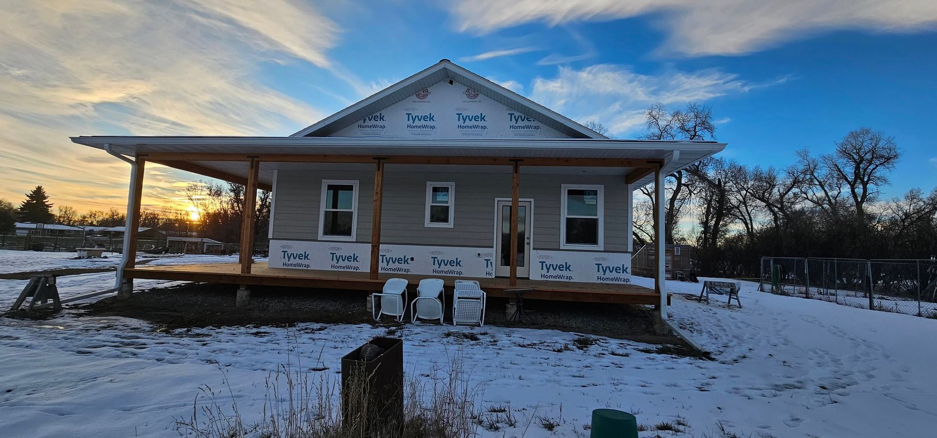 A house under construction with light grey siding and a large front porch sits in a snowy landscape at sunset.