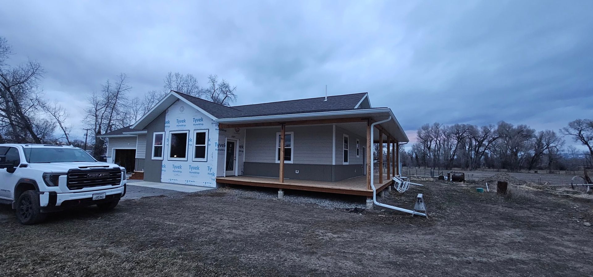 A new house under construction with light-colored siding, a wrap-around porch, and a white truck parked in the foreground.