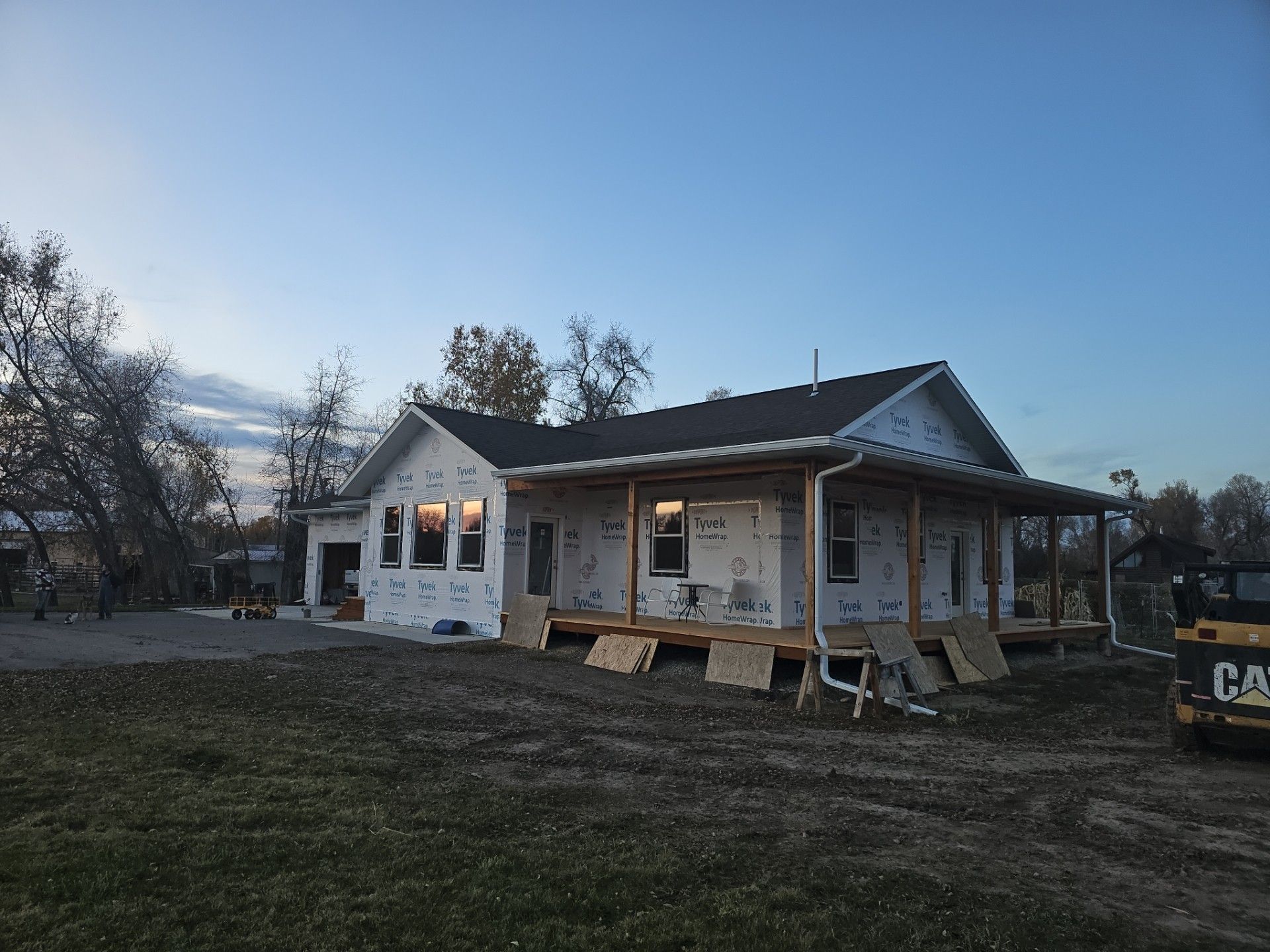 A house under construction with white exterior sheathing, a dark roof, and a wooden porch, set in a field under a blue sky.