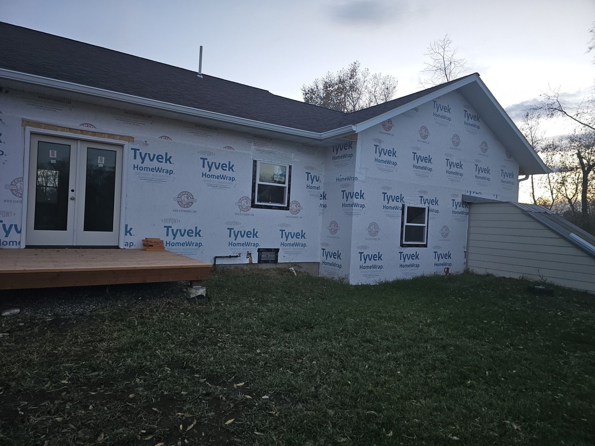 A house exterior under construction with white Tyvek house wrap, a wooden deck, and a basement stairwell.