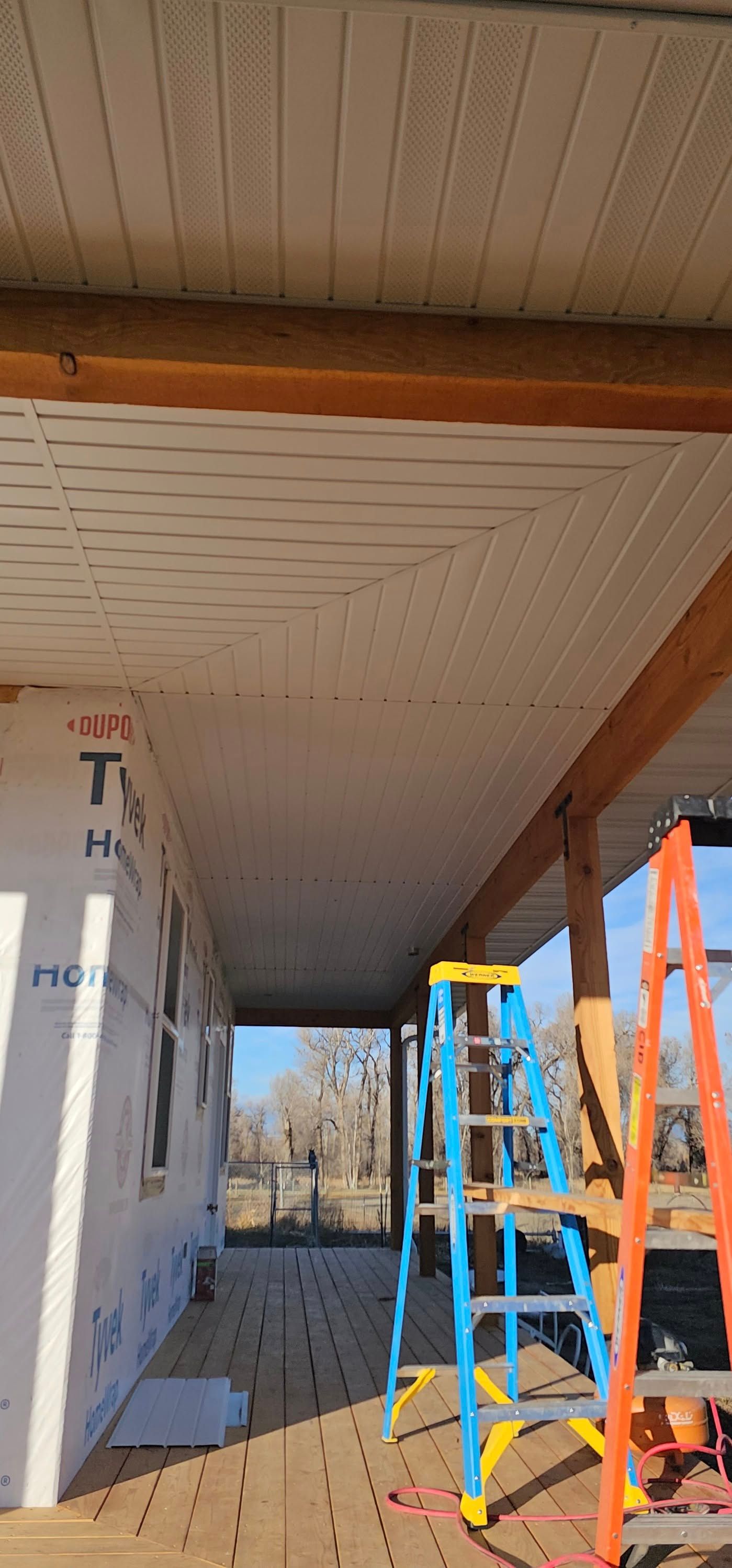A white vinyl soffit ceiling under construction on a porch, with wooden framing, two ladders, and a blue sky in the distance.