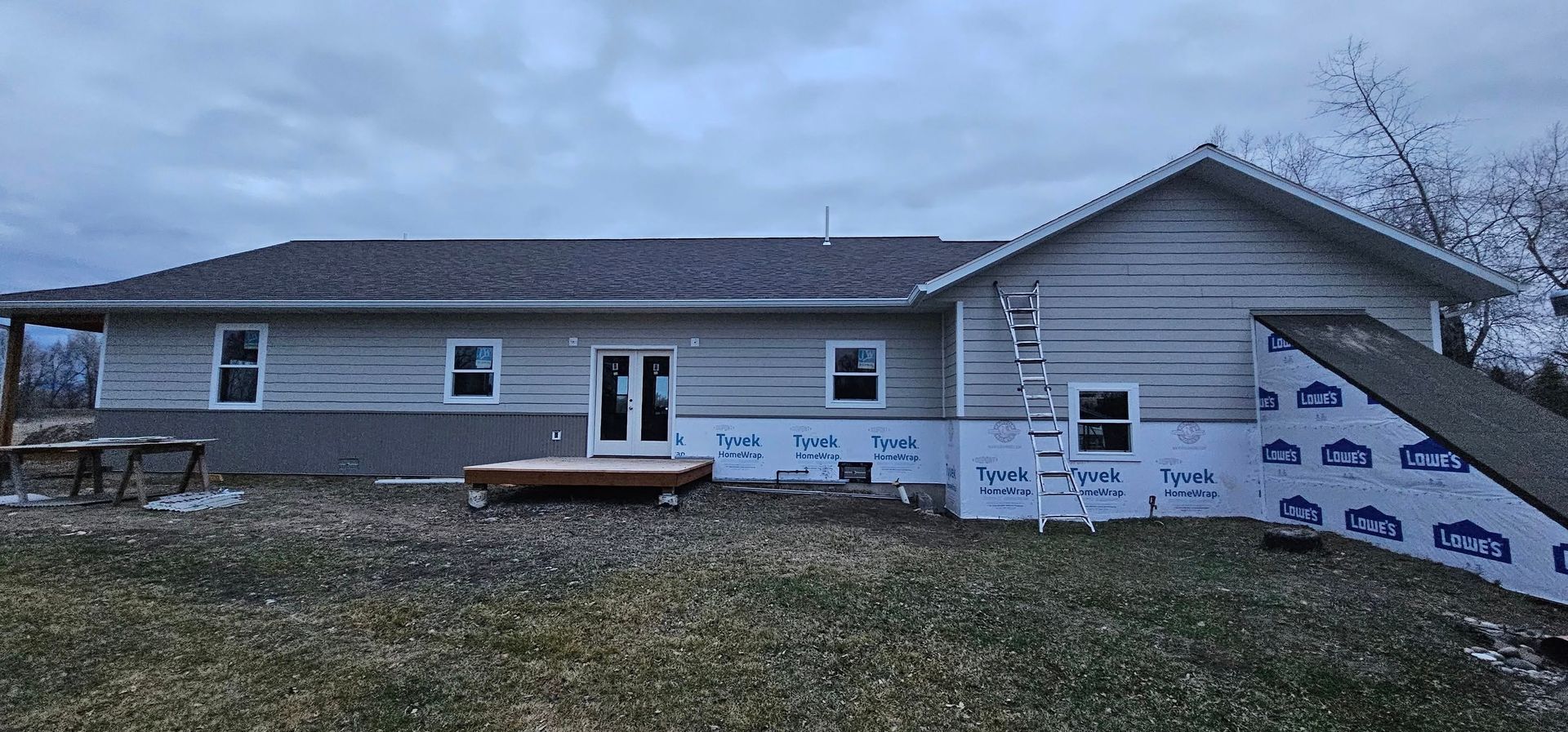 A ranch-style house under construction with light gray siding, exposed white house wrap, and a ladder leaning against it.