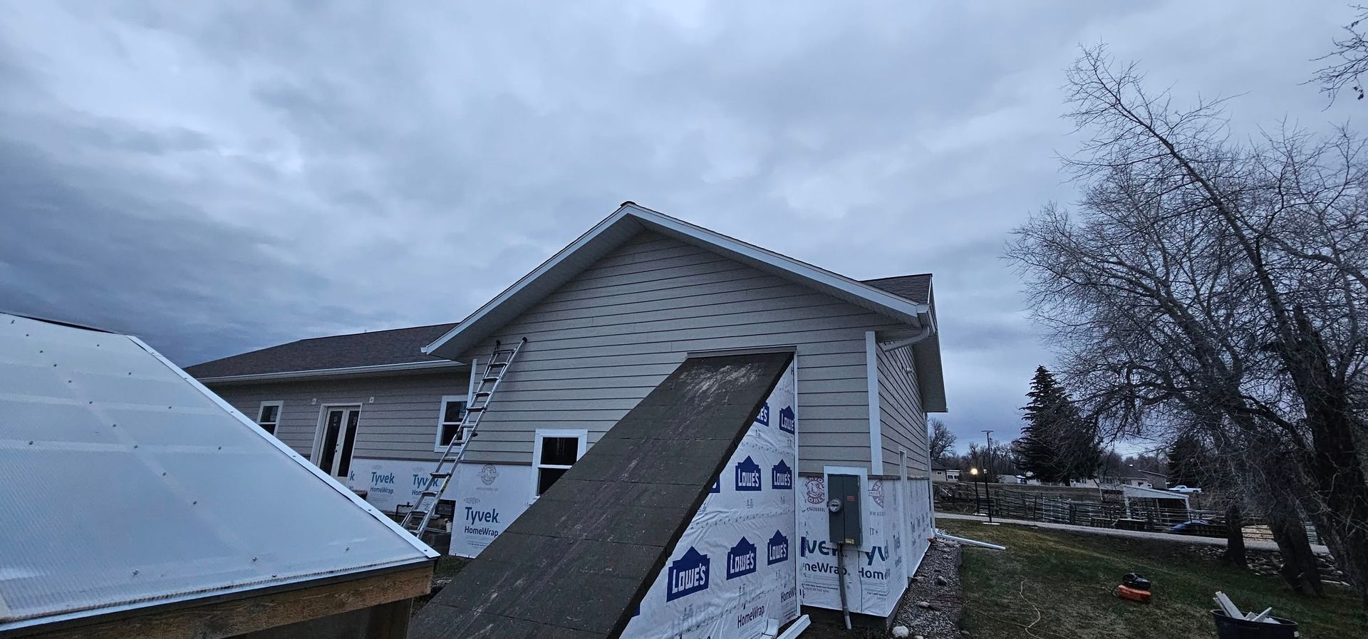 A beige house under construction with white siding, a metal roof, and blue protective wrap on a cloudy day.