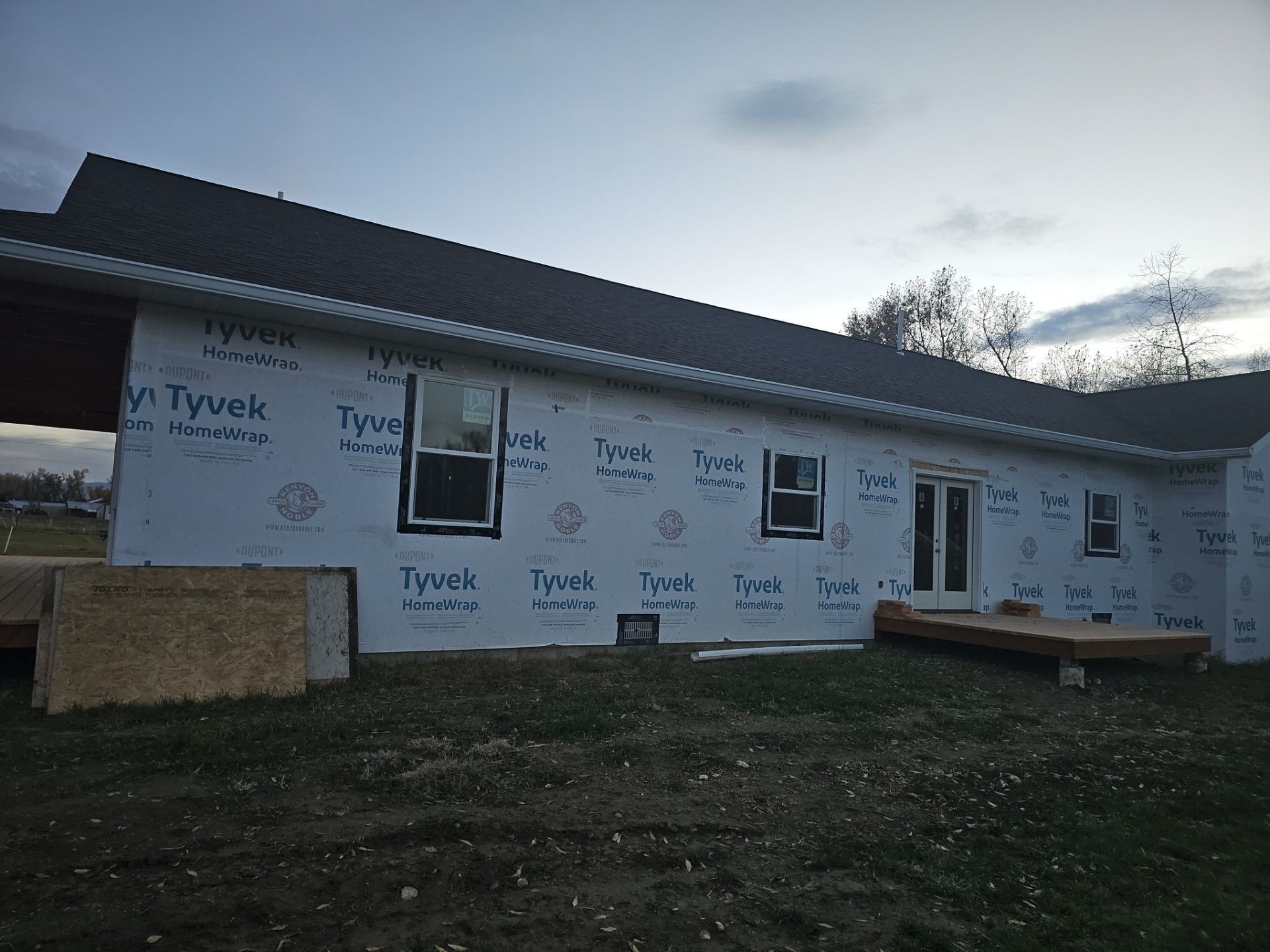 A house under construction with white Tyvek siding, a dark roof, two windows, and a set of exterior doors.