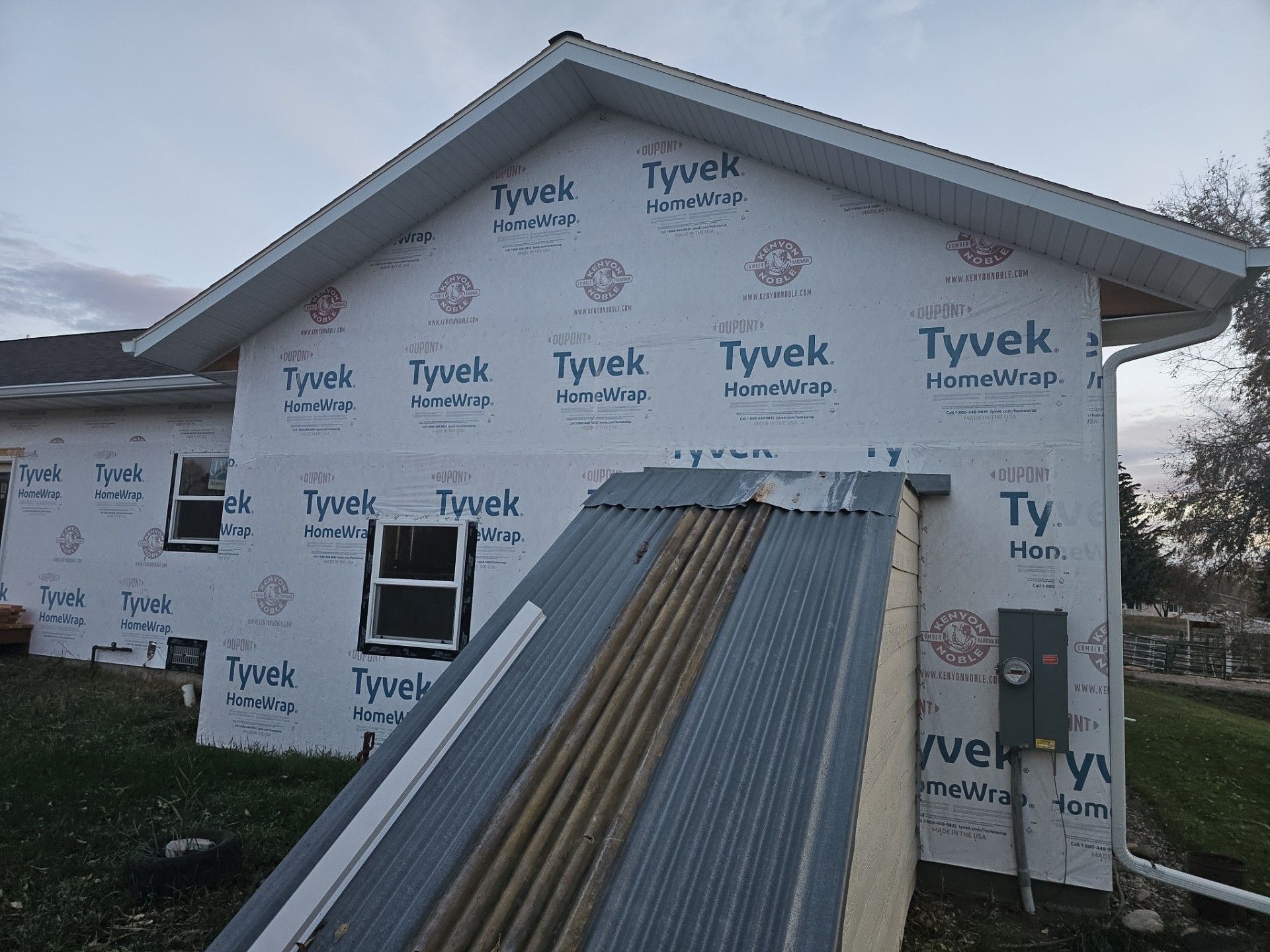 A house exterior covered in white Tyvek building wrap with a sloped metal cellar door entrance in the foreground.