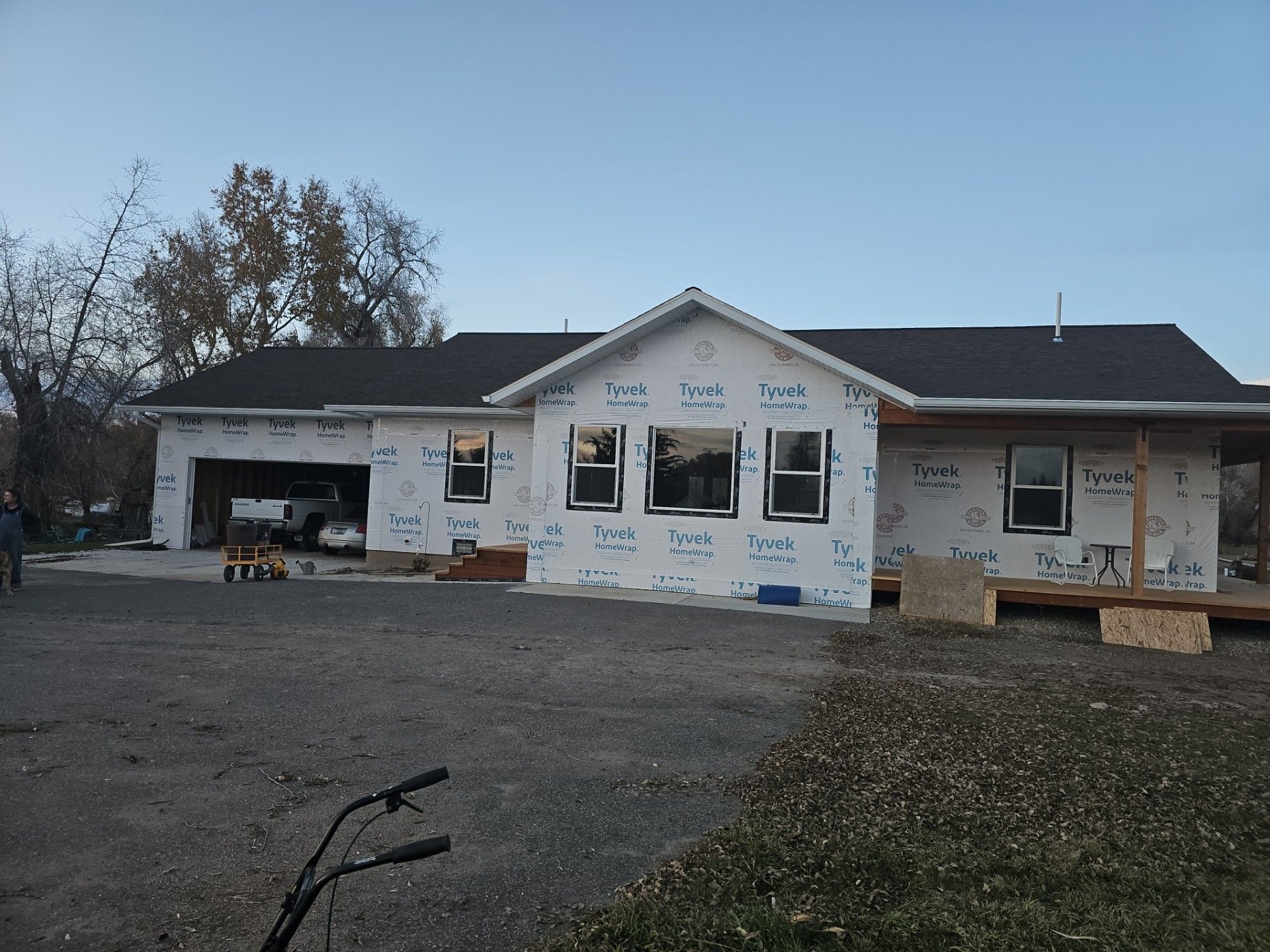 A single-story house under construction with white Tyvek house wrap, dark roofing, and open garage, set on a gravel lot.