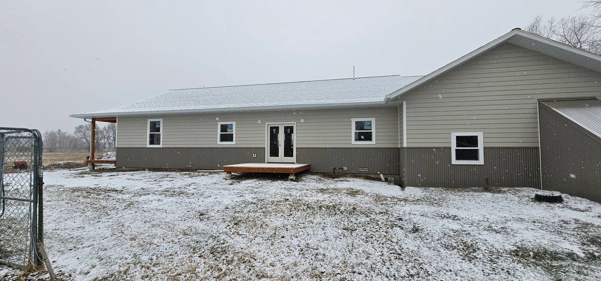 A long, one-story house with beige and grey siding, a small wooden deck, and light snow on the roof and ground.