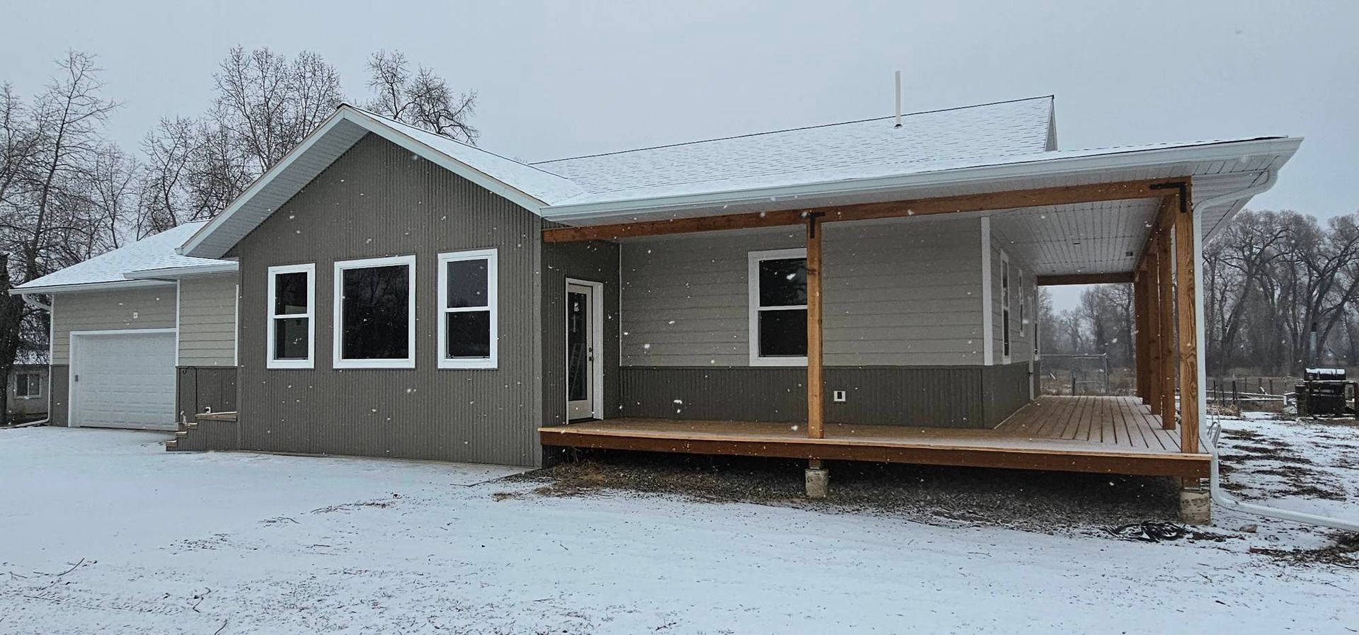 A modern, light-gray and dark-gray house with a large wrap-around porch stands on a snow-covered, rural landscape.