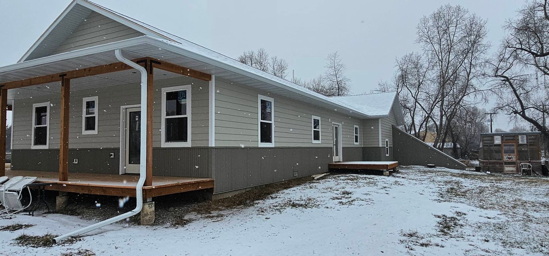 A one-story house with light gray horizontal siding over dark gray vertical paneling, a wooden porch, and snow-covered ground.