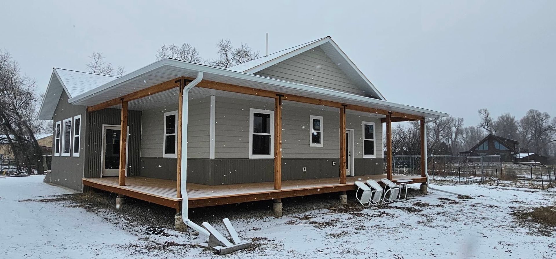 A one-story house with gray siding and a wrap-around wooden porch sits in a snow-covered yard.