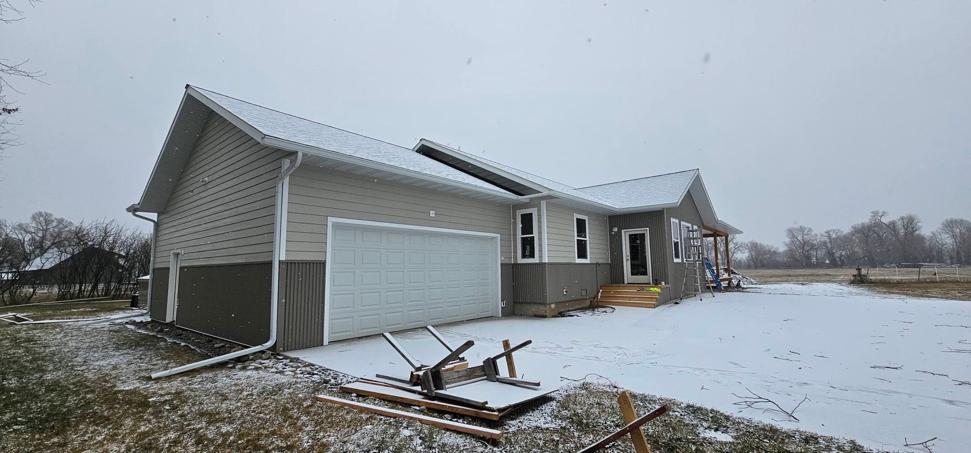 A beige house with a white garage door and dark foundation sits on a snow-covered yard under a cloudy sky.