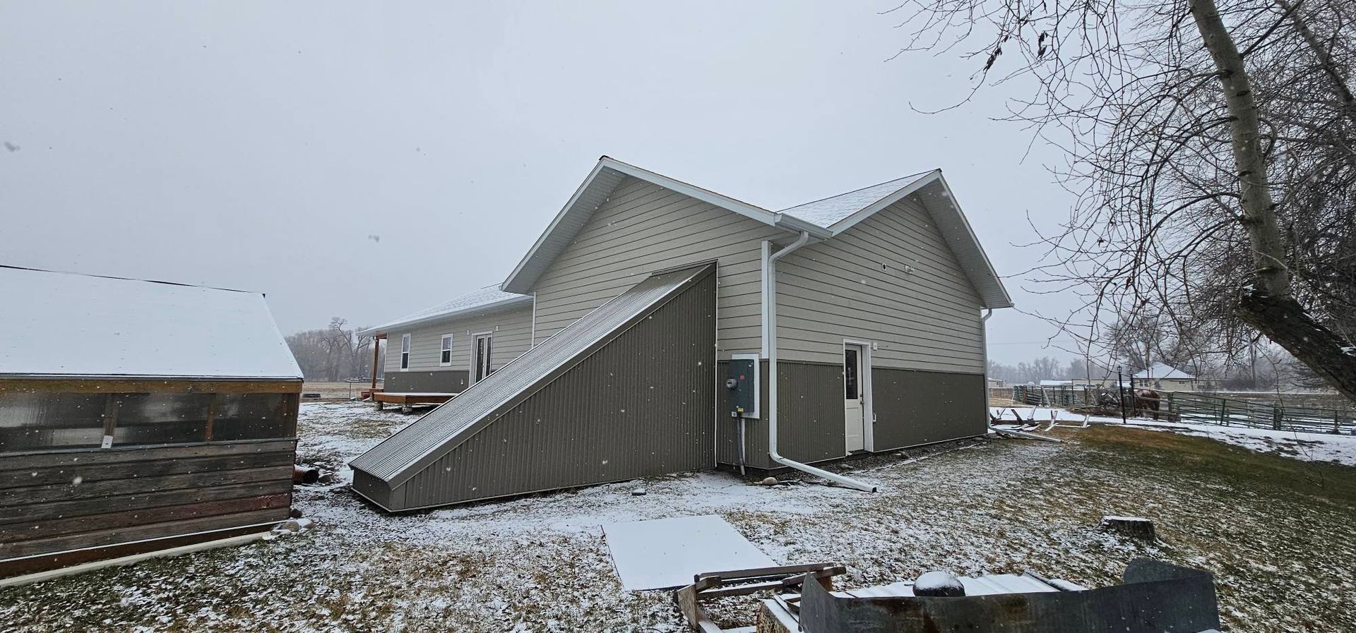 A light-colored house with a large, slanted wooden structure attached to its side, set in a light dusting of snow.