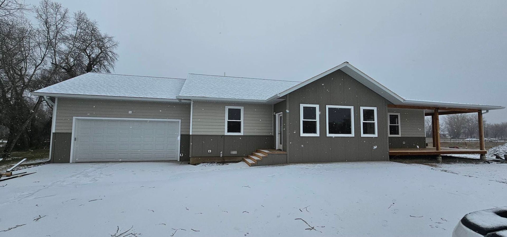 A modern single-story house with a two-car garage, grey siding, and a wooden porch, set in a snowy landscape.