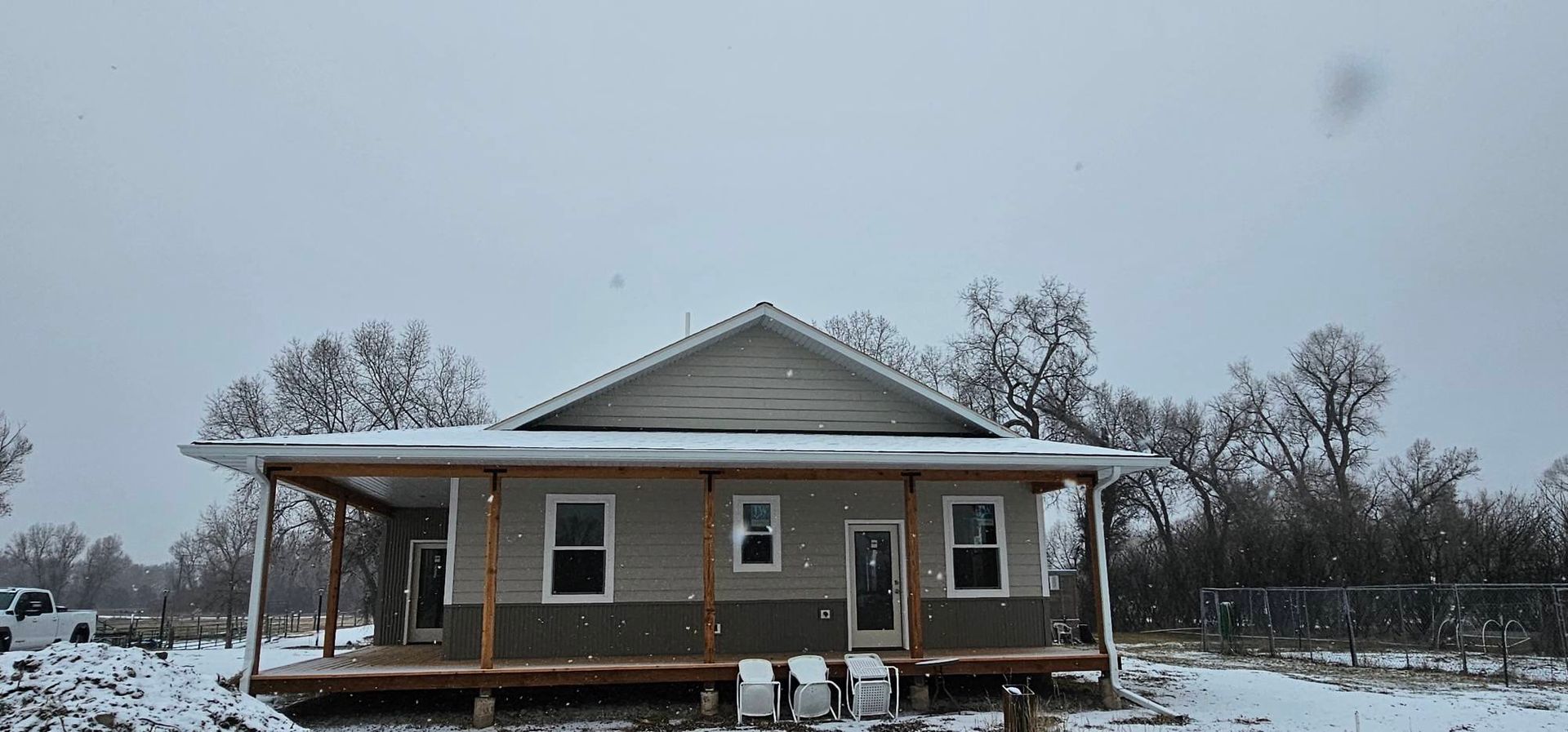 A one-story house with a large front porch and two-toned gray siding, surrounded by a snowy yard under a gray sky.