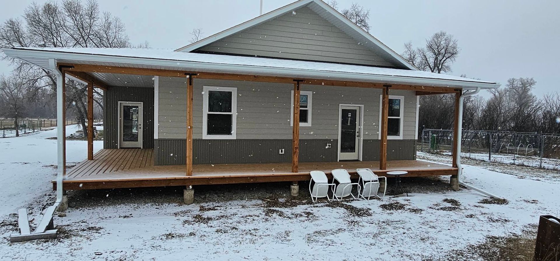 A light gray house with a wooden wrap-around porch and metal roof covered in a dusting of light snow.