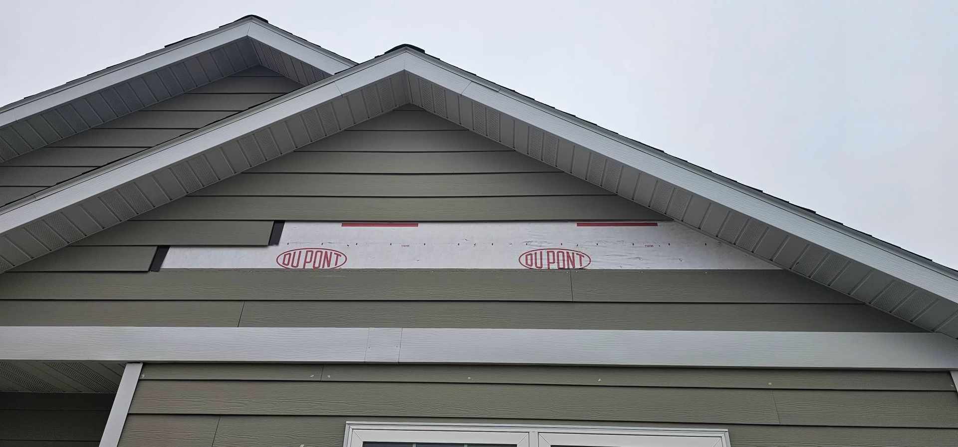 Gray horizontal siding on a gable-roofed house with a missing section revealing white moisture barrier underlayment.