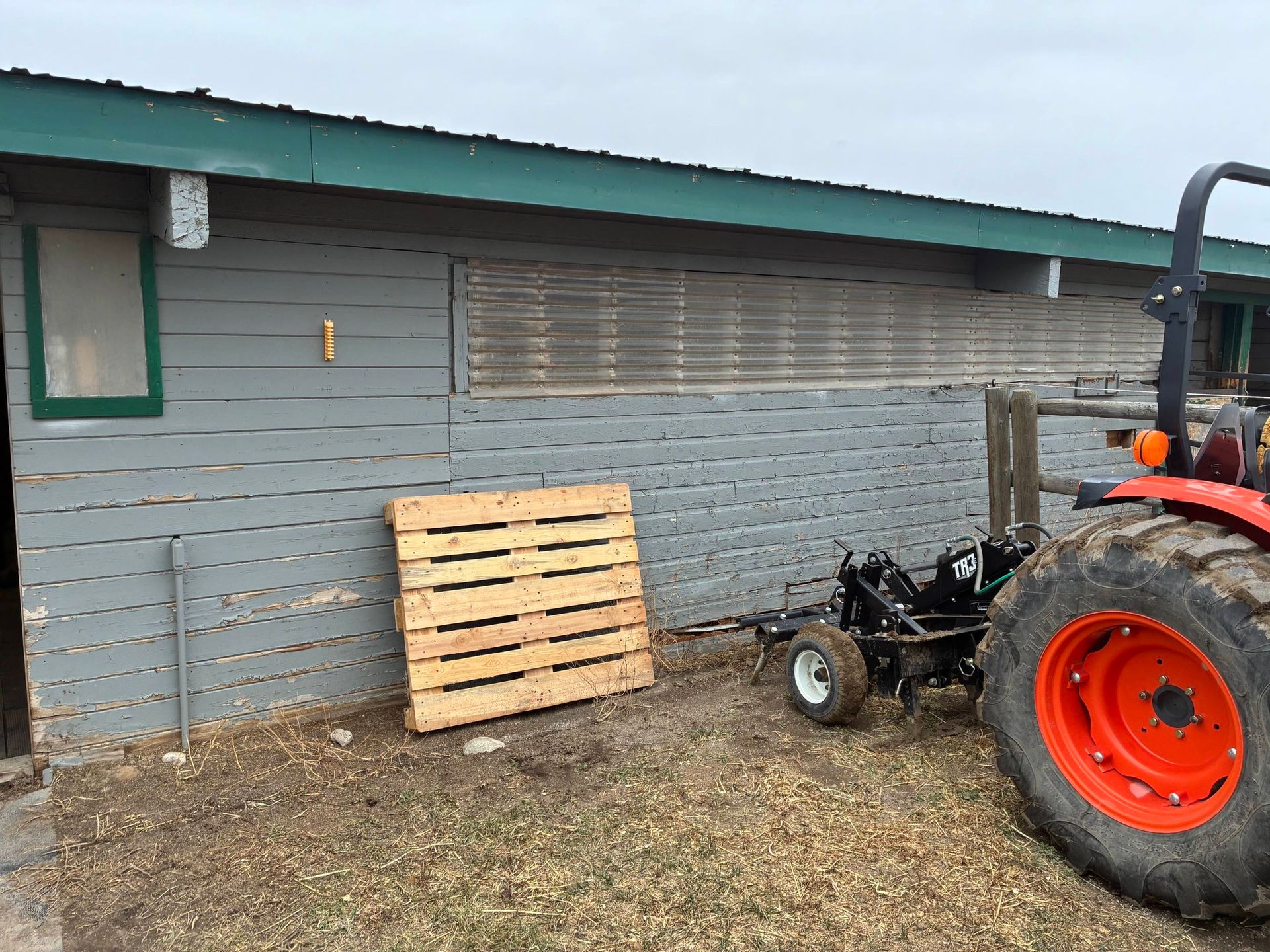 A tractor sits parked next to a gray wooden building with a horizontal slatted vent and a wooden pallet leaning on the wall.