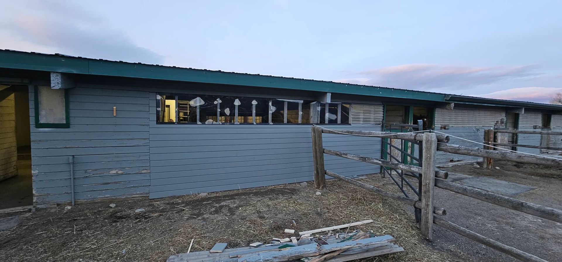 A grey, single-story building with a green roof, windows, and an adjacent wooden fence on a gravel lot at twilight.
