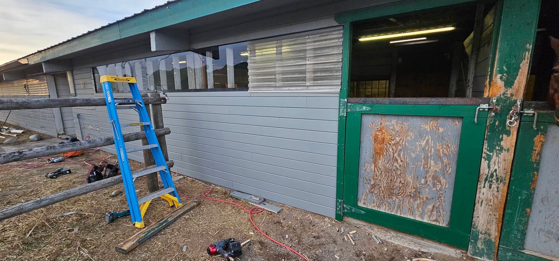 A partially renovated stable exterior with grey siding, green trim, and a blue ladder set up for construction.