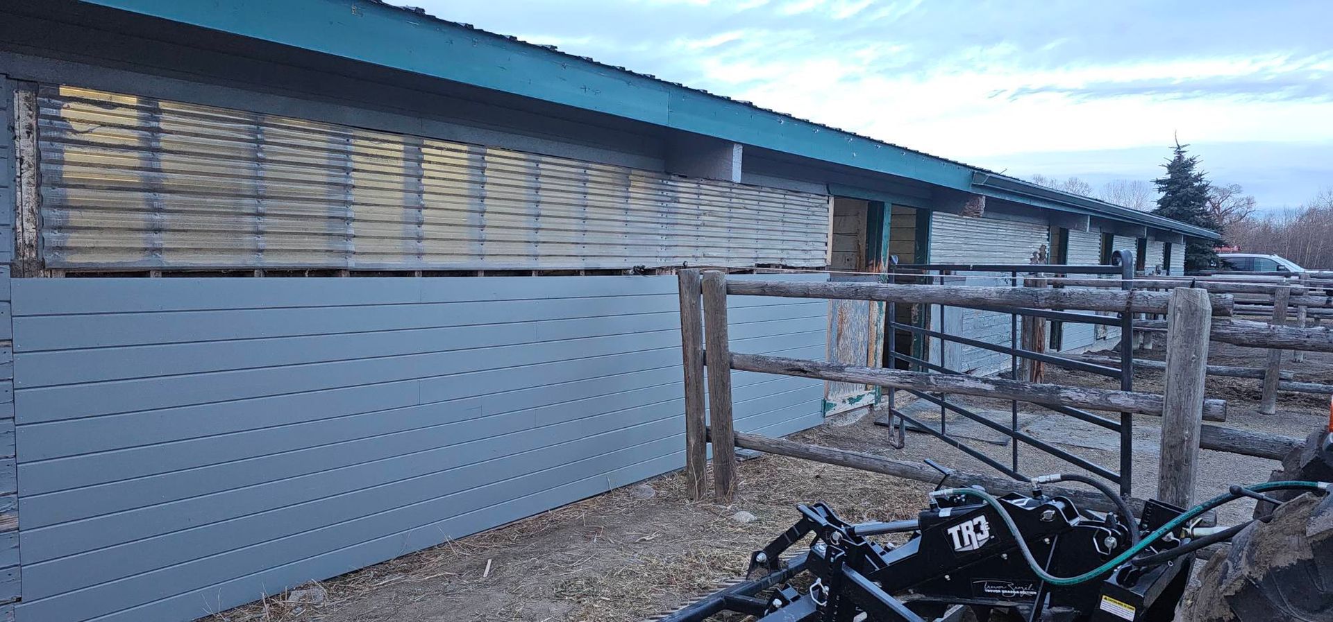 A long, gray-sided horse stable with slat vents and a blue roof, situated next to a wooden fence and farm equipment.