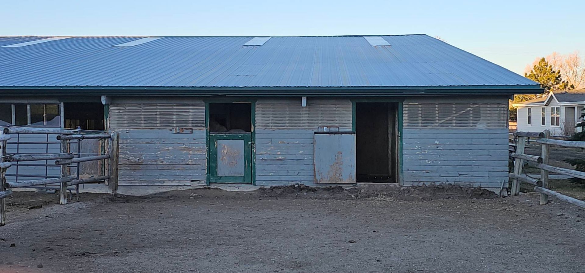 A long, single-story stable with a blue metal roof, grey cinder block walls, and open horse stalls in a dirt paddock.